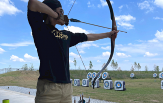 Person shooting a recurve bow with bright blue skies in the background and archery targets behind them in Bentonville, Arkansas